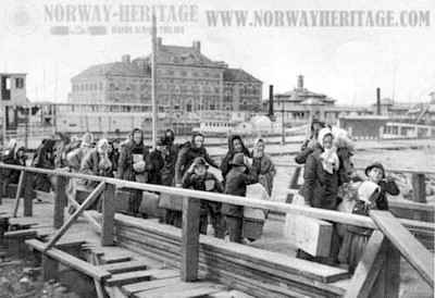 Emigrants coming up the board-walk from the barge which had taken them off the steamship company docks and transported them to Ellis Island. The hospital in the background. In the middle of the picture the ferry boat running to New York